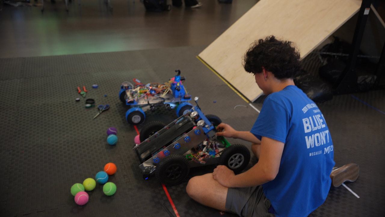 A student kneeling beside two robots on the course, next to a plywood ramp