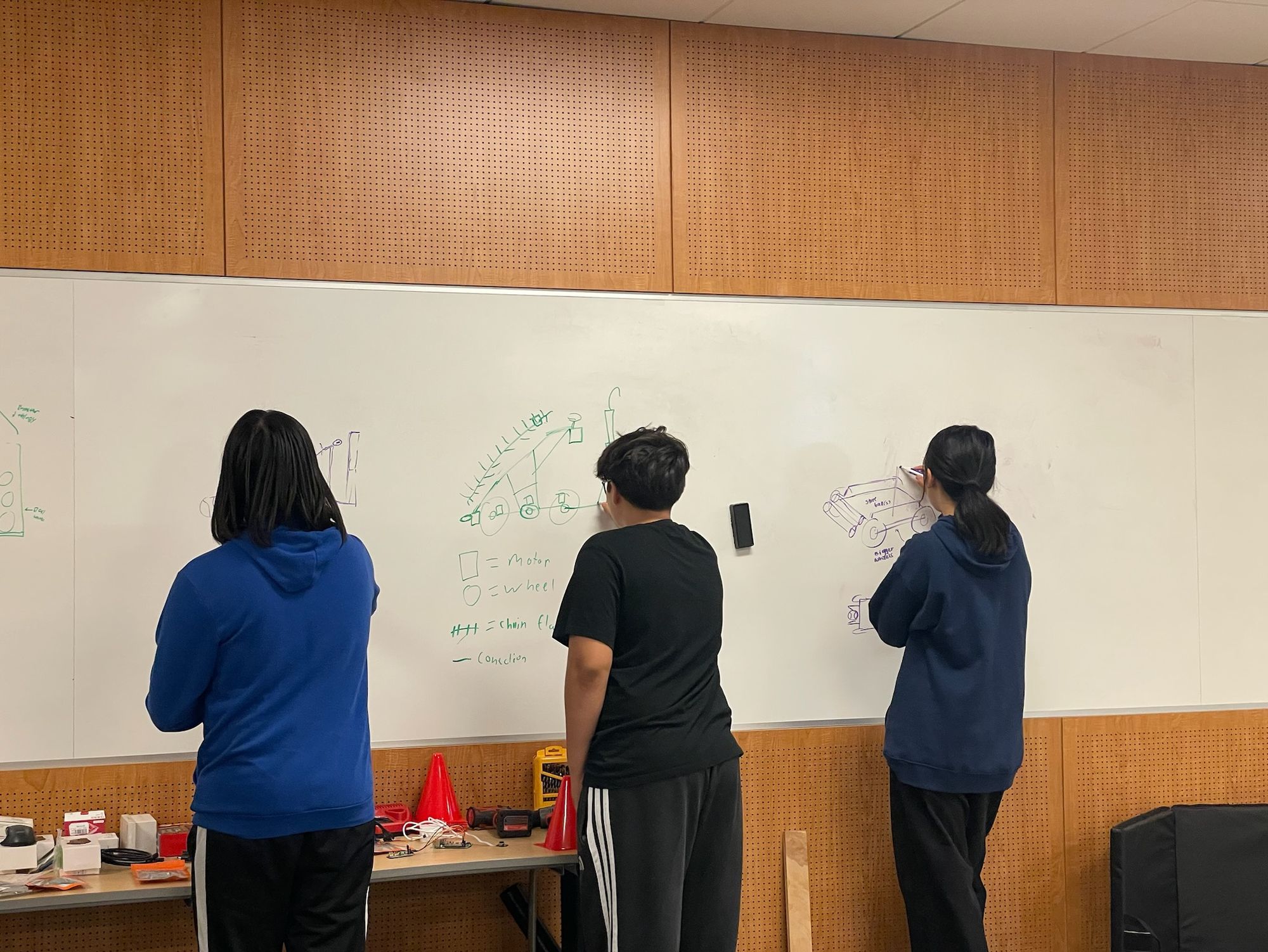Three students sketching robot designs on a whiteboard